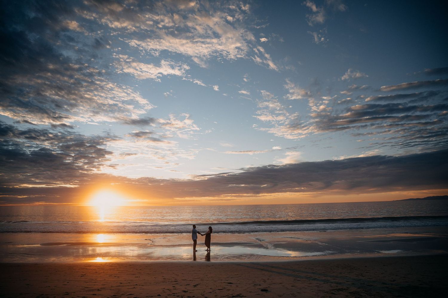 Two silhouettes stand on a beach at sunset with dramatic clouds and reflective waves along the shoreline.