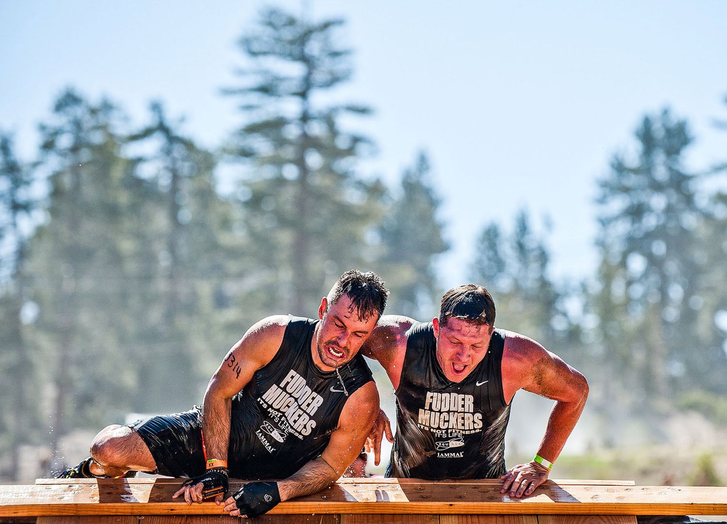 Two athletes in black tank tops performing synchronized push-ups on wooden platform during outdoor training event.