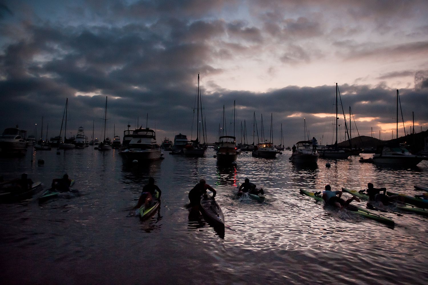 Silhouetted boats anchored in a marina at sunset with dramatic cloudy skies reflecting on the dark water.