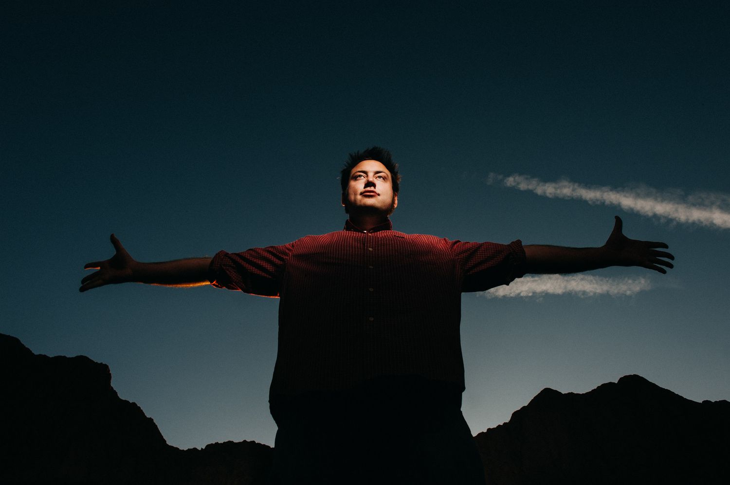 Silhouette of person with outstretched arms against dramatic mountain landscape and dark blue sky at dusk.