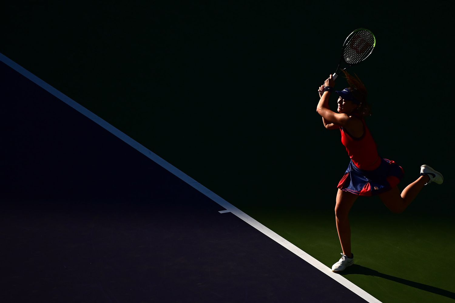 Tennis player serves under dramatic lighting on a tennis court casting long shadows during a match.