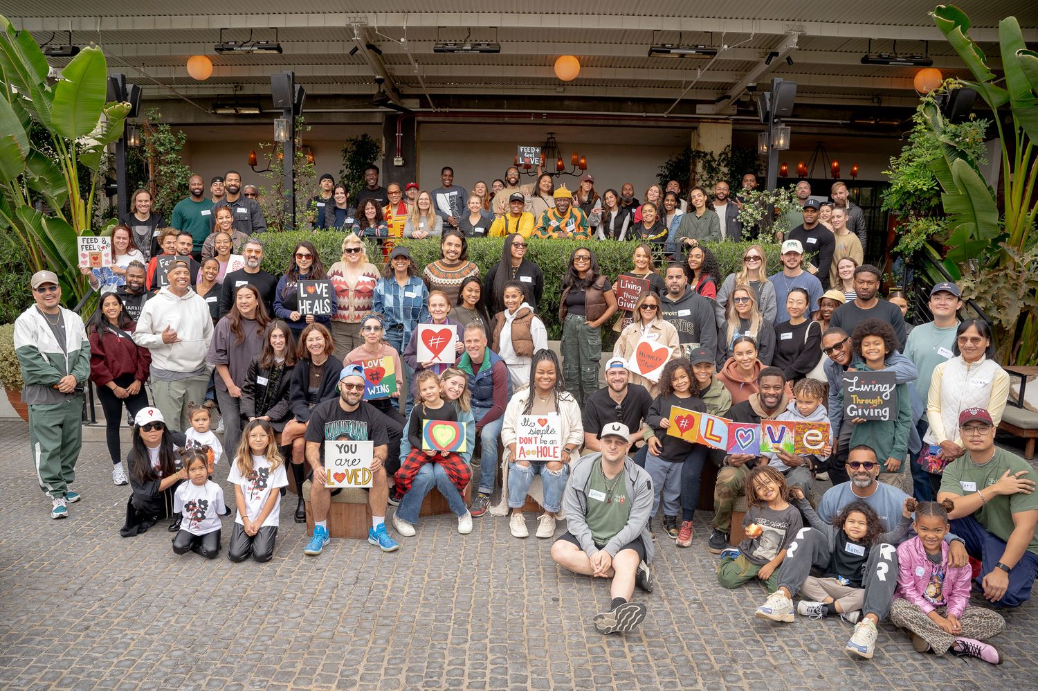 Large group of people gathered outdoors for a group photo in front of a building entrance with tropical plants.