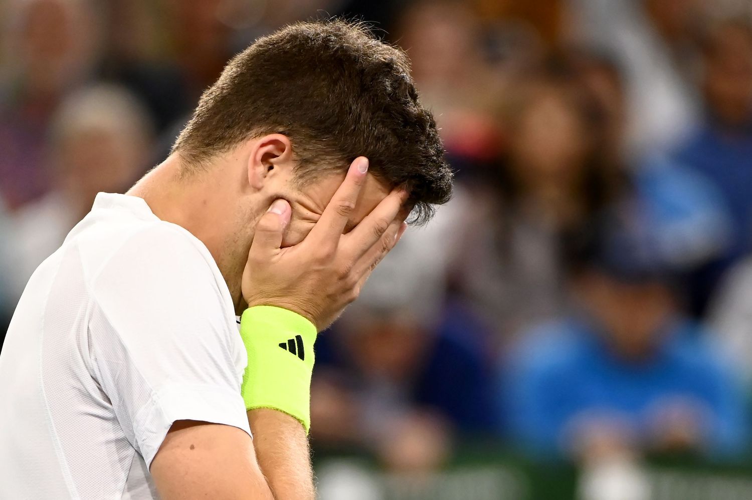 A tennis player in white attire covers face with hand in disappointment during a match.