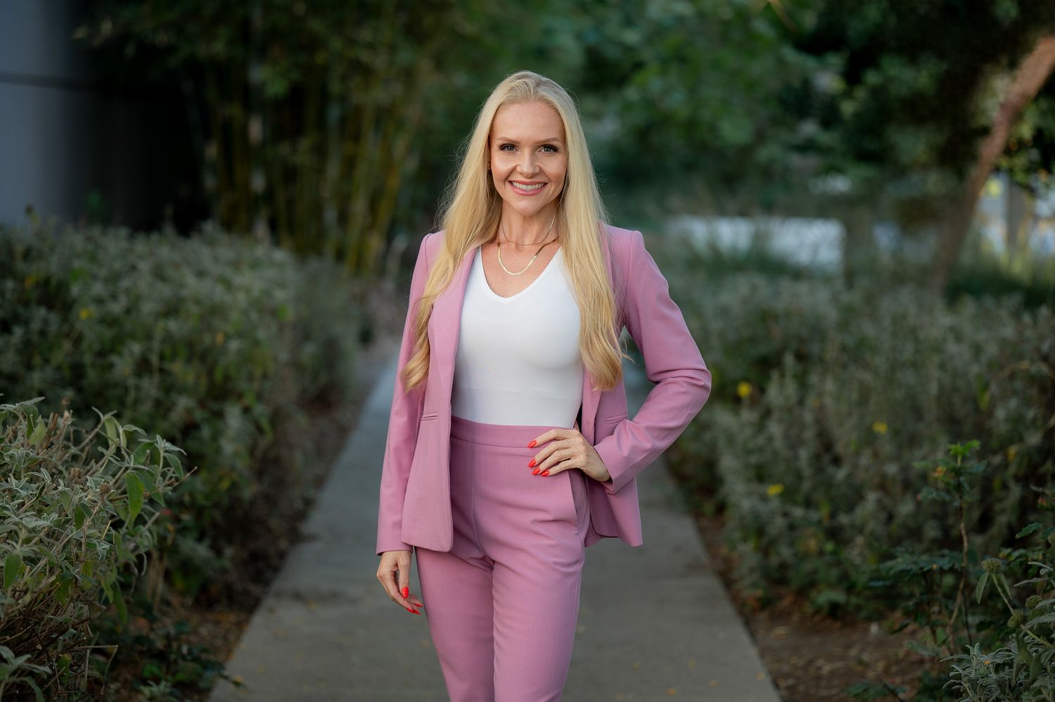 Professional portrait of person in pink business suit and white top standing on outdoor pathway surrounded by greenery.