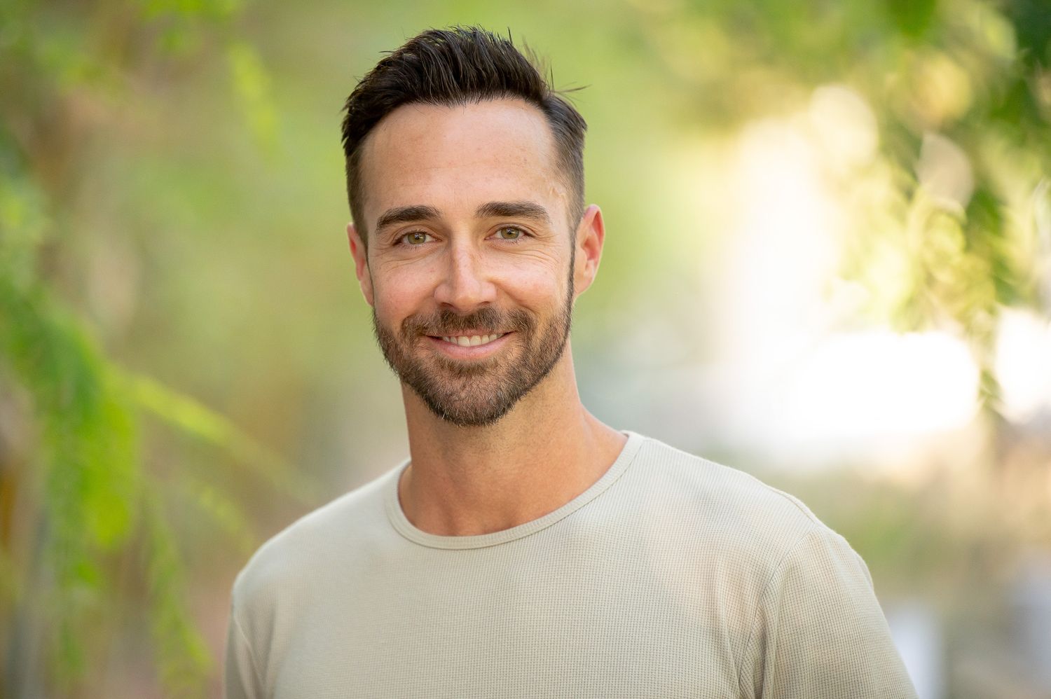 A person with facial hair wearing a light gray t-shirt smiles at the camera with soft natural lighting and foliage in background.