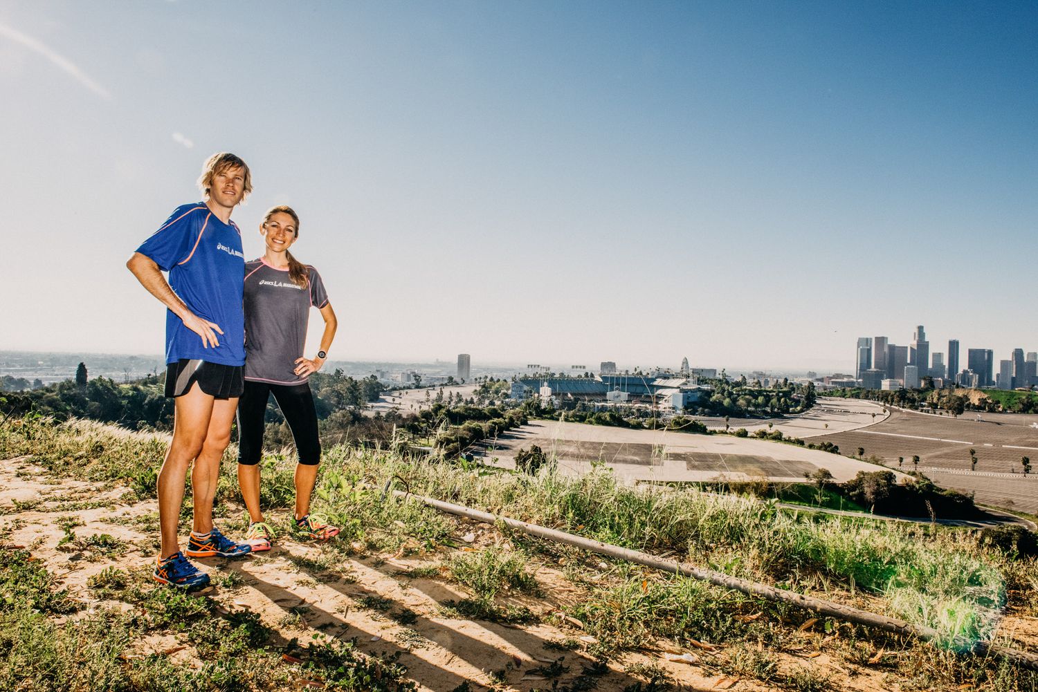 Two people in athletic wear stand on a hilltop overlooking the Los Angeles city skyline on a clear sunny day.