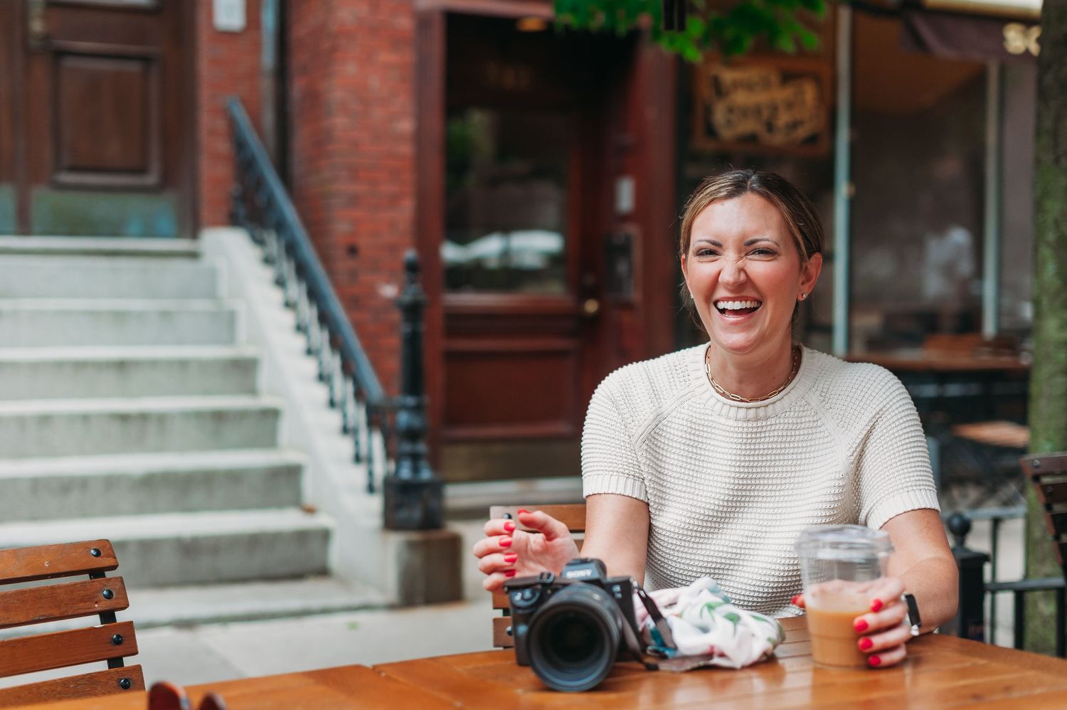 Someone sits at an outdoor cafe table with a camera and beverages, enjoying a casual afternoon on a city street.