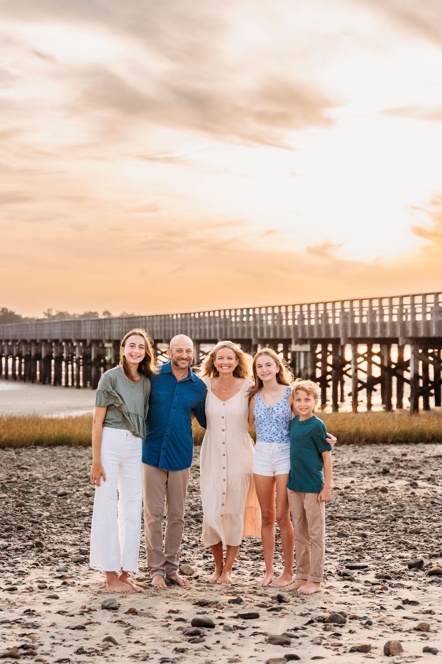 A loving family group poses together on a sandy beach at sunset with a long wooden pier in the background.