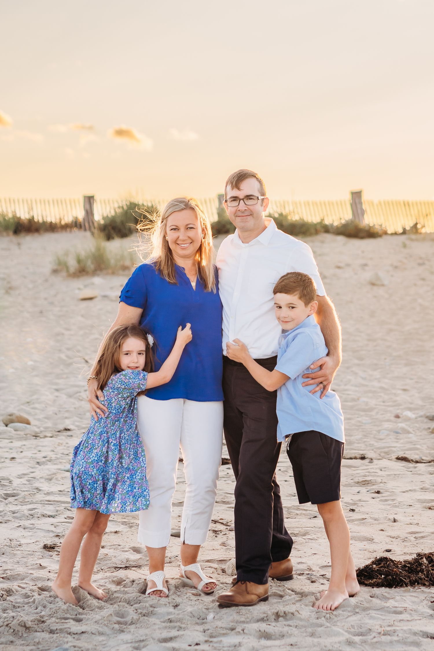 A family of four stands together on a sandy beach at sunset wearing coordinated blue and white outfits.