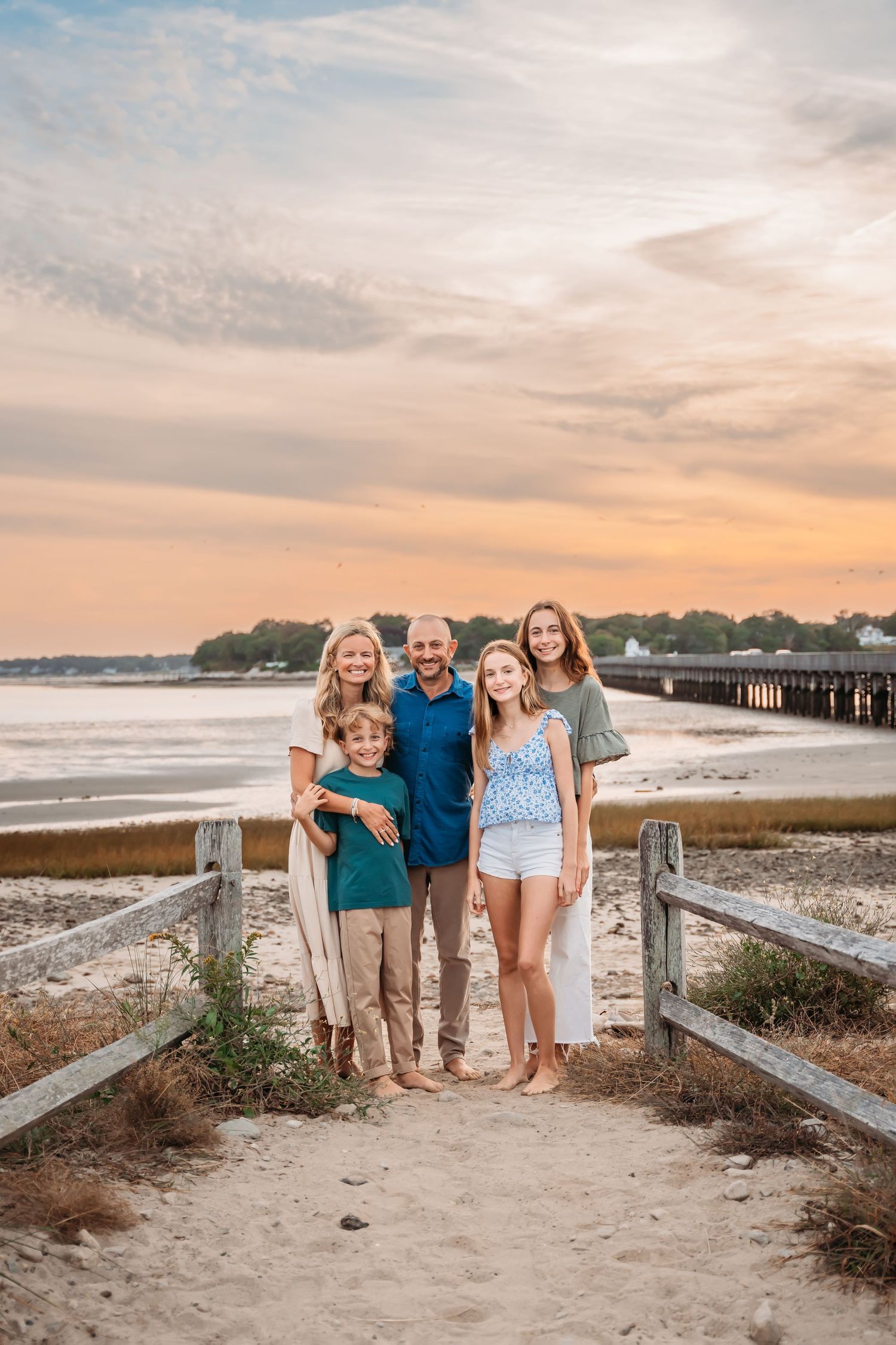 A family stands together on a sandy beach path with a pier and sunset ocean view in the background during golden hour.