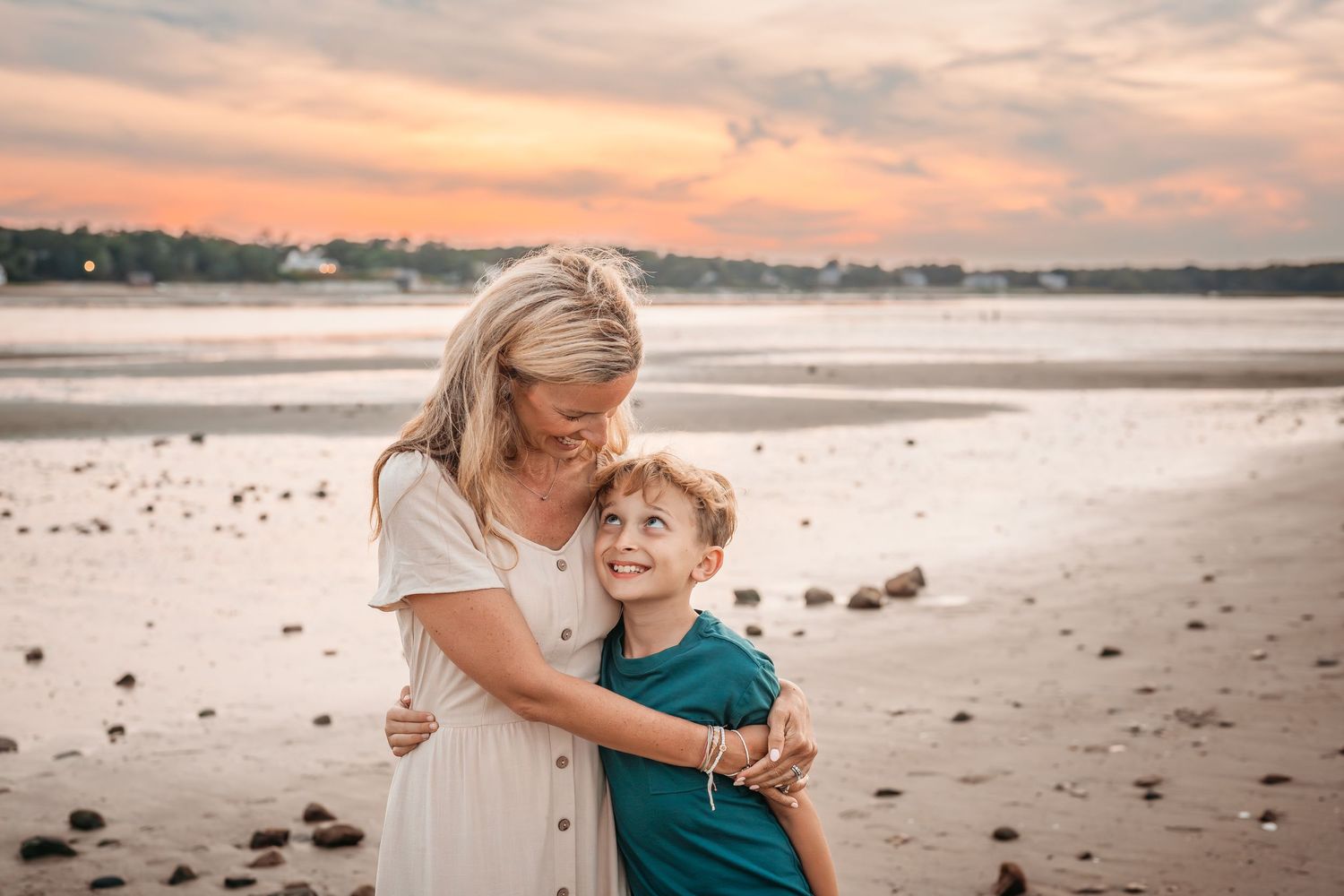 A parent and child share a tender moment embracing on a beach at sunset with orange sky and calm waters in the background.