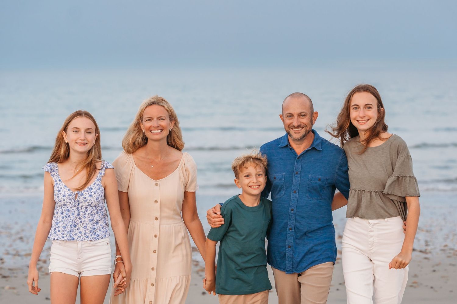 A family of five posing together at the beach during sunset, wearing casual attire in neutral and blue tones.