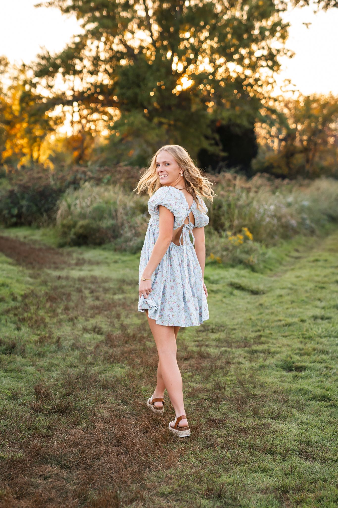 Young girl in light blue floral dress walking in a grassy meadow at sunset with golden light.