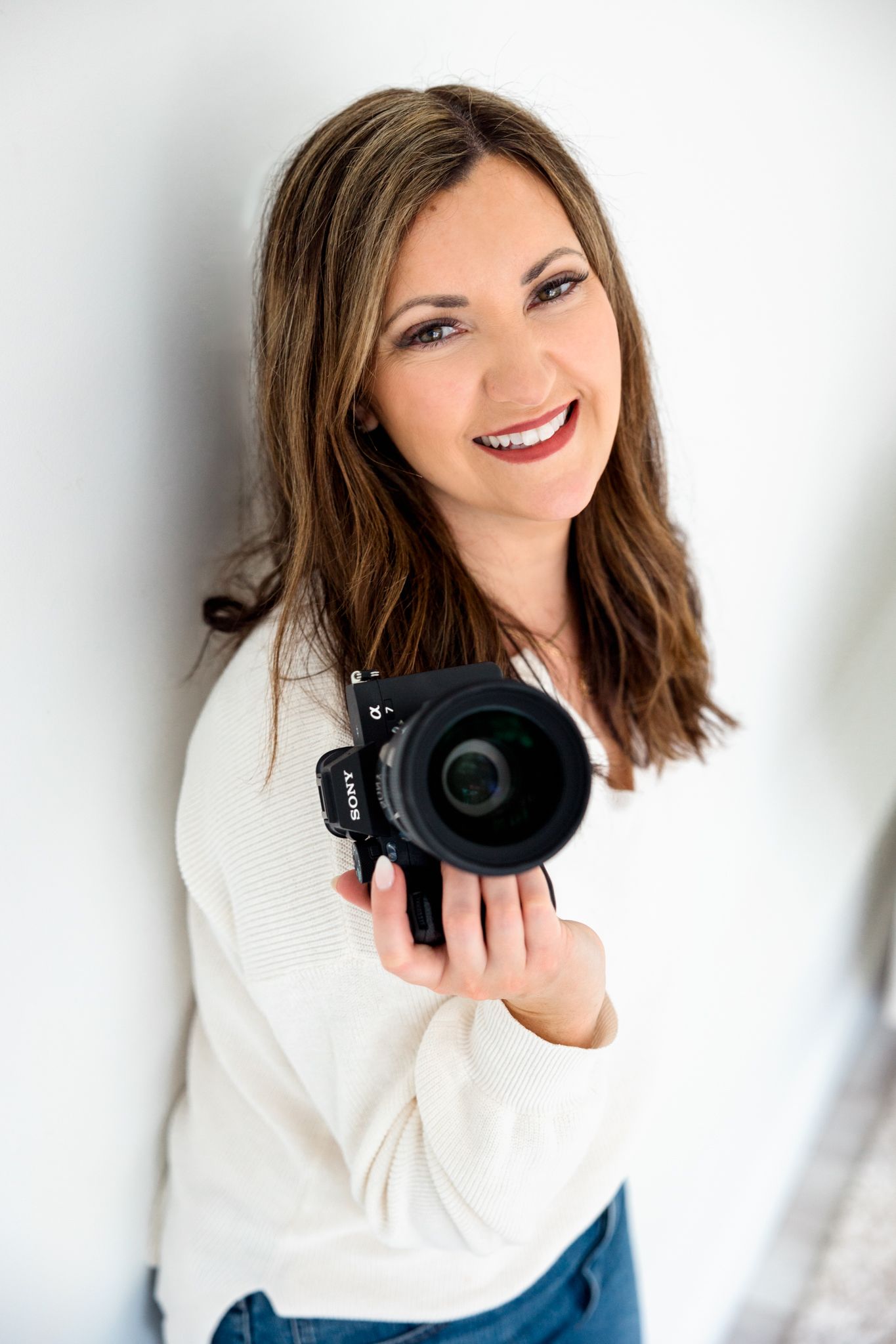 Professional photographer in white sweater holds camera while posing against white background.