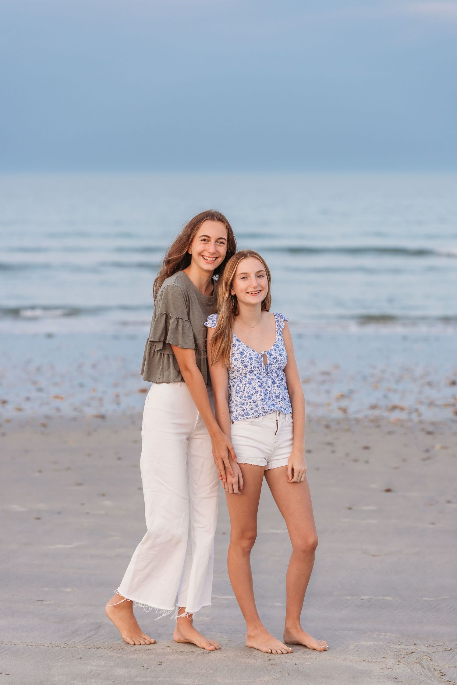 Two people in casual summer outfits standing together barefoot on a beach at sunset.