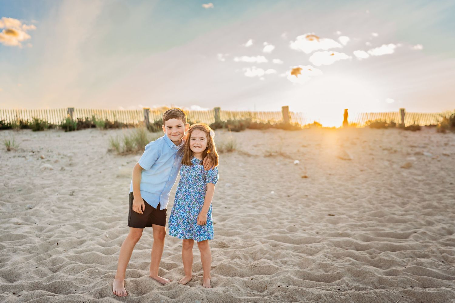 Two people stand together on a sandy beach at sunset with a city skyline visible in the distance.