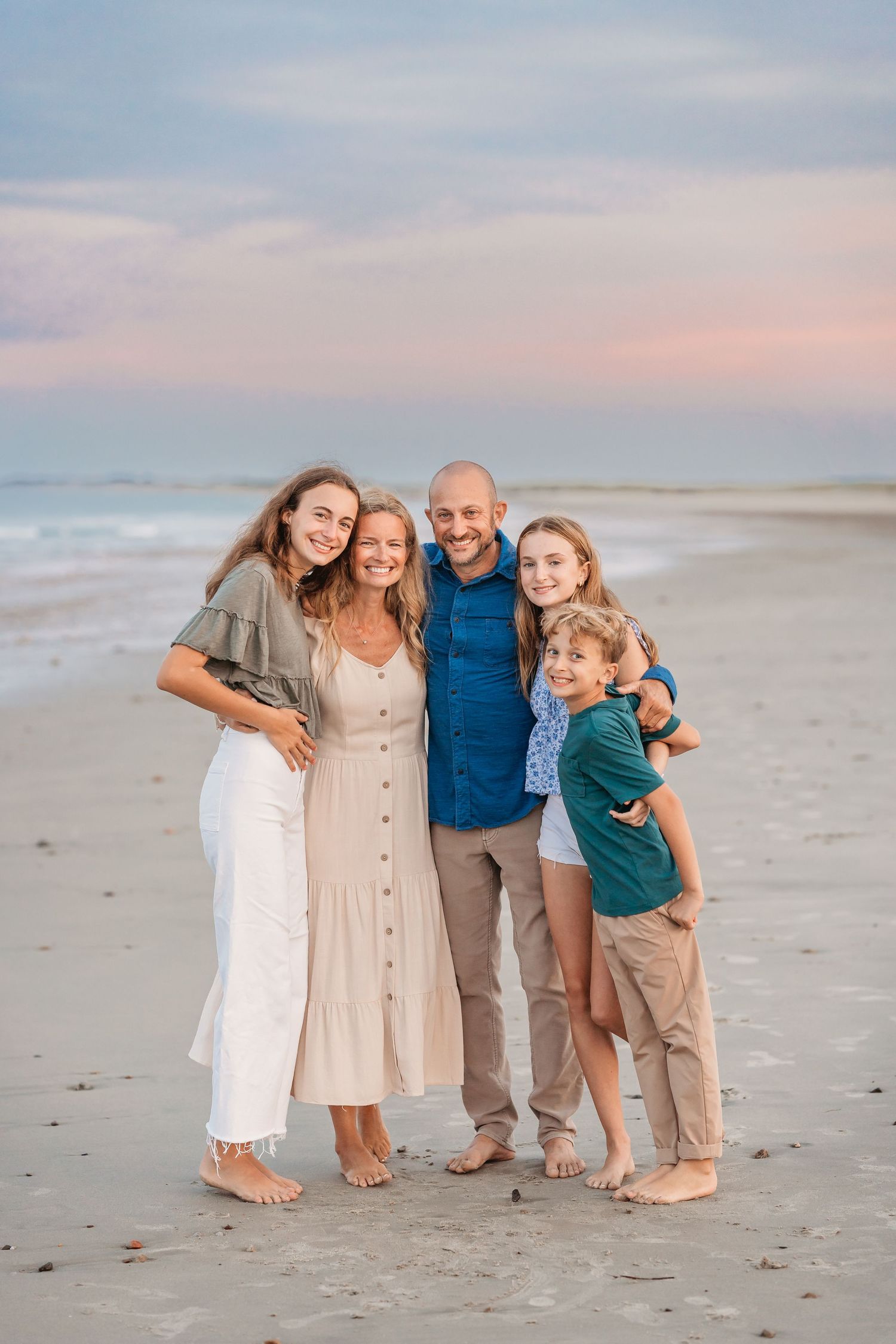 A joyful family of five standing together barefoot on a peaceful beach during sunset with soft pink sky.