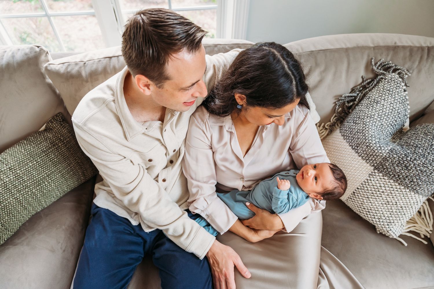 Couple sitting on grey couch while tenderly holding their newborn baby.