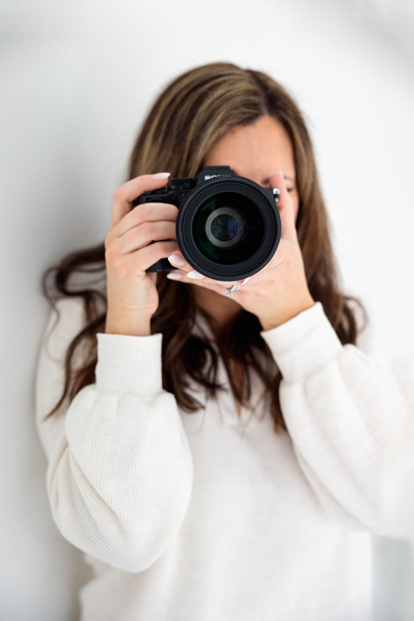 Photographer in white sweater holds professional camera in front of face against white background.