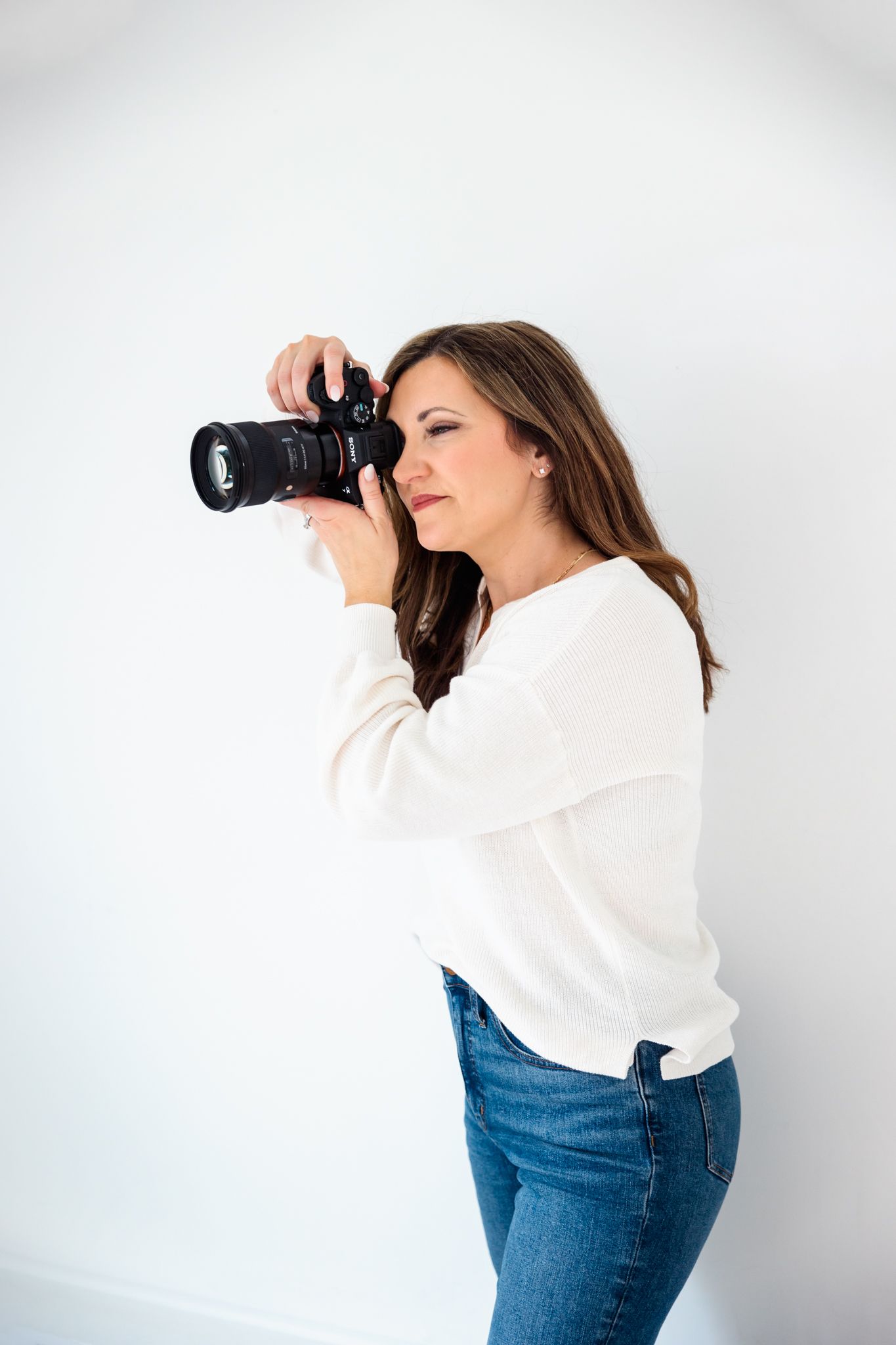 Photographer in casual attire takes photos against white studio backdrop.