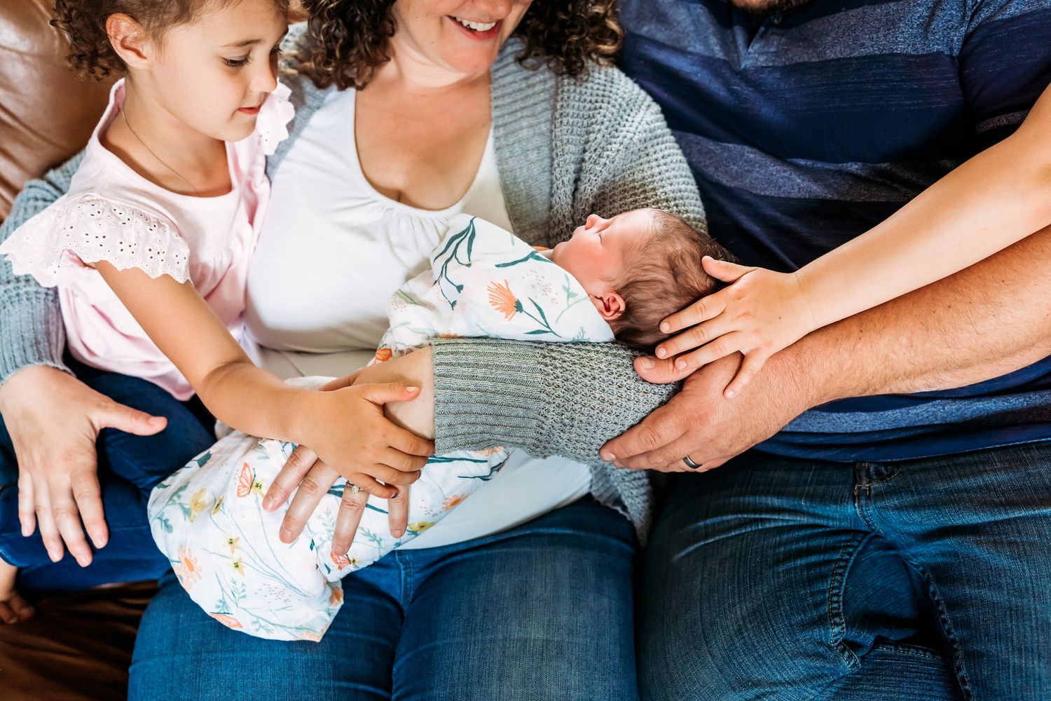 Family sitting closely together on blue couch while holding and admiring newborn baby wrapped in gray blanket.