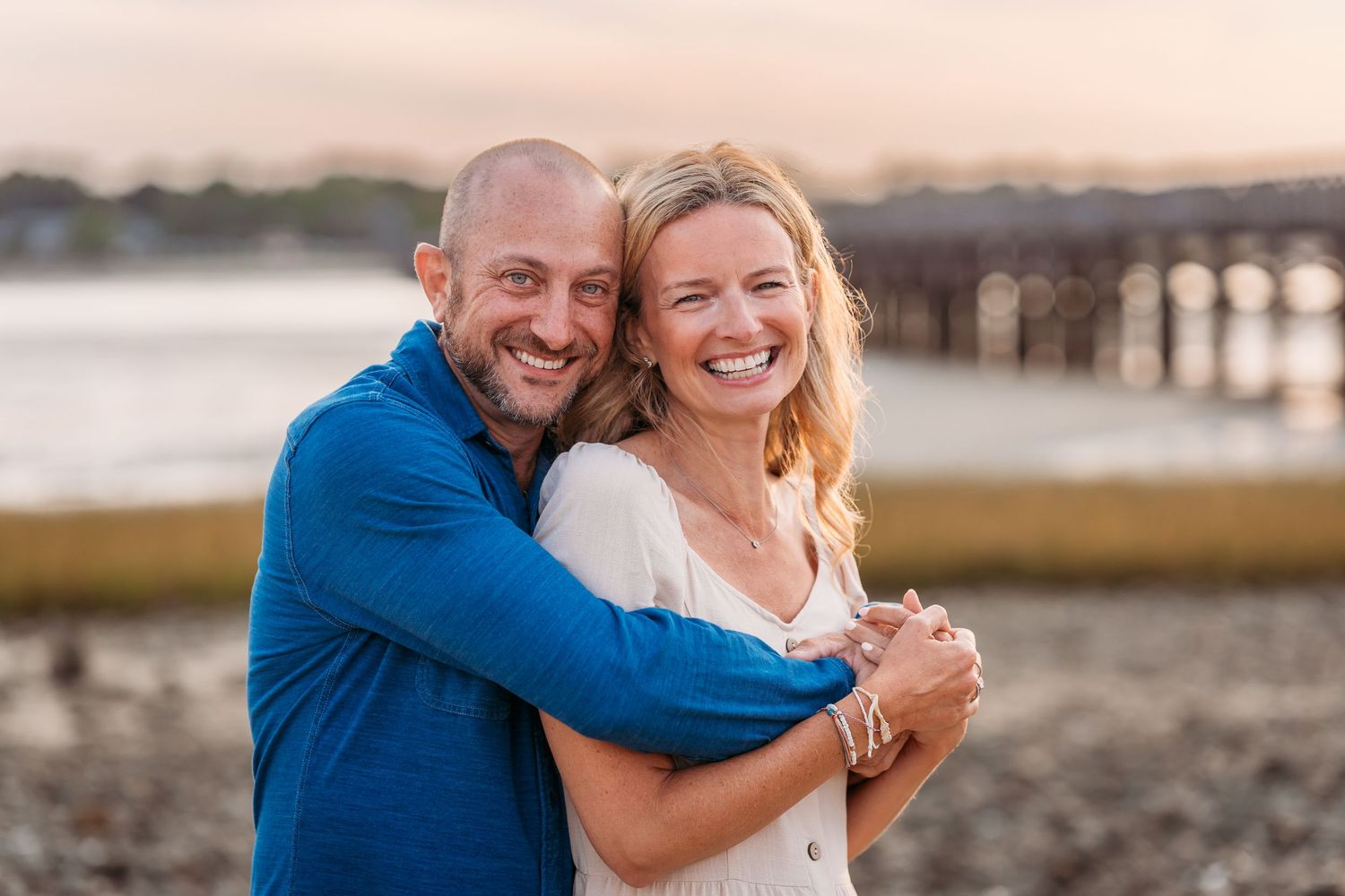 Happy couple embraces and laughs together on beach at sunset with wooden pier in background.