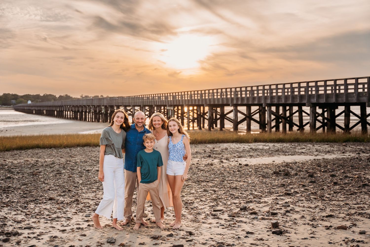 A group stands together on a beach at sunset with a long wooden pier stretching into the distance.