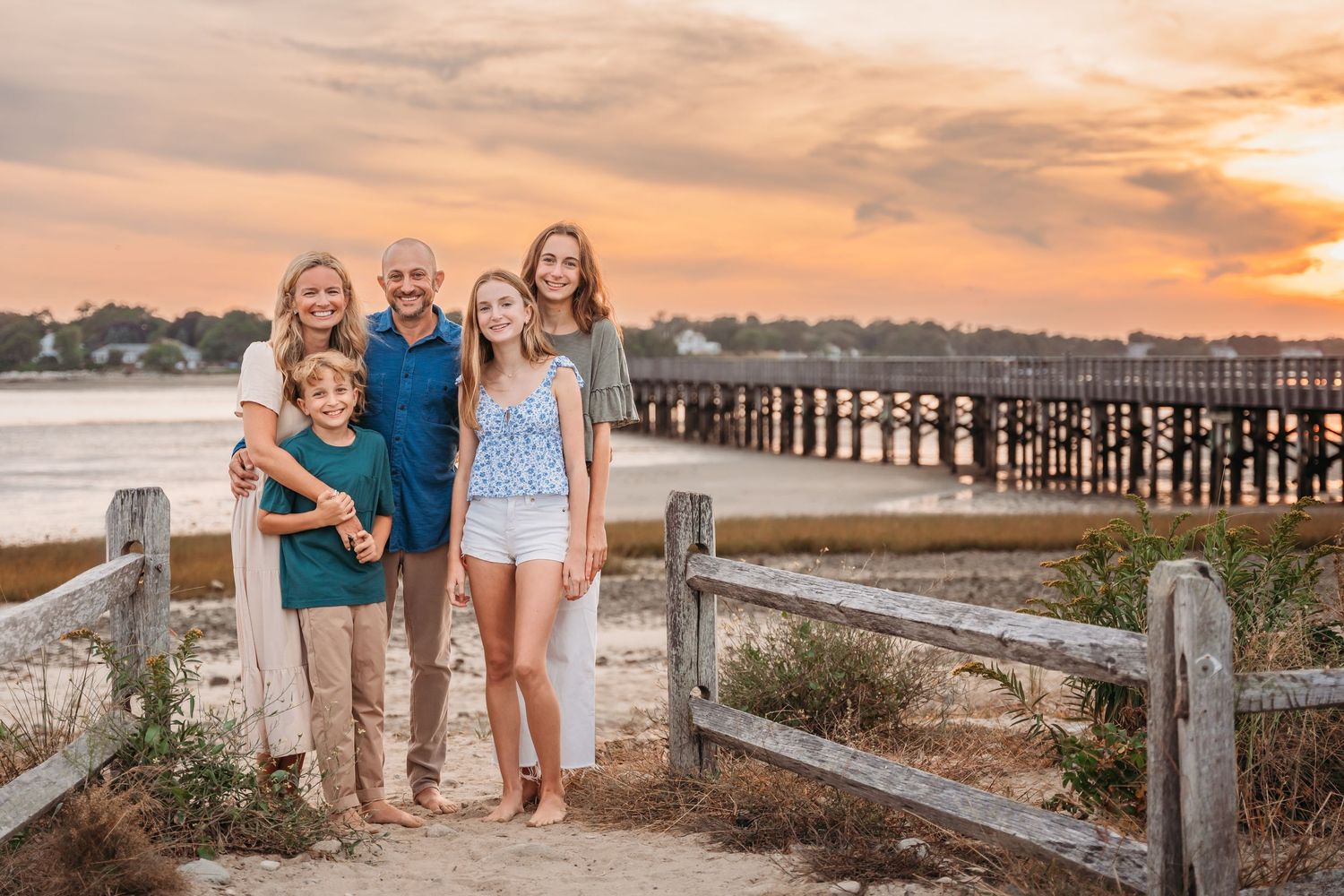 A group poses together at sunset near a wooden pier and beach fence with warm golden light in the background.