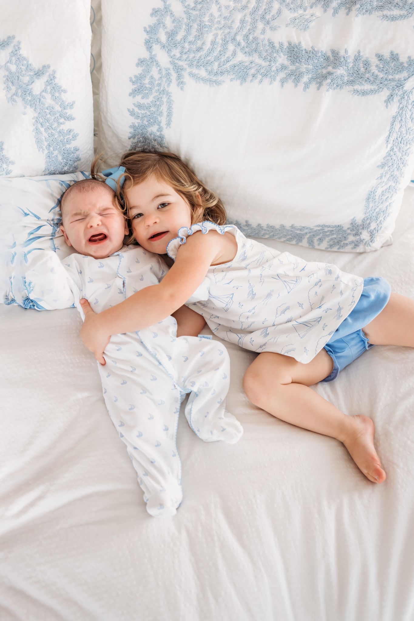 Two young children cuddle and laugh together on a blue and white patterned bedspread.