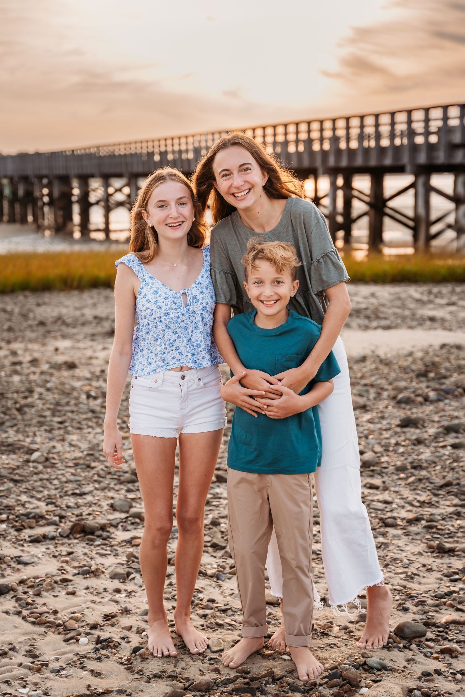 Family members stand barefoot on a beach at sunset with a wooden pier in the background.
