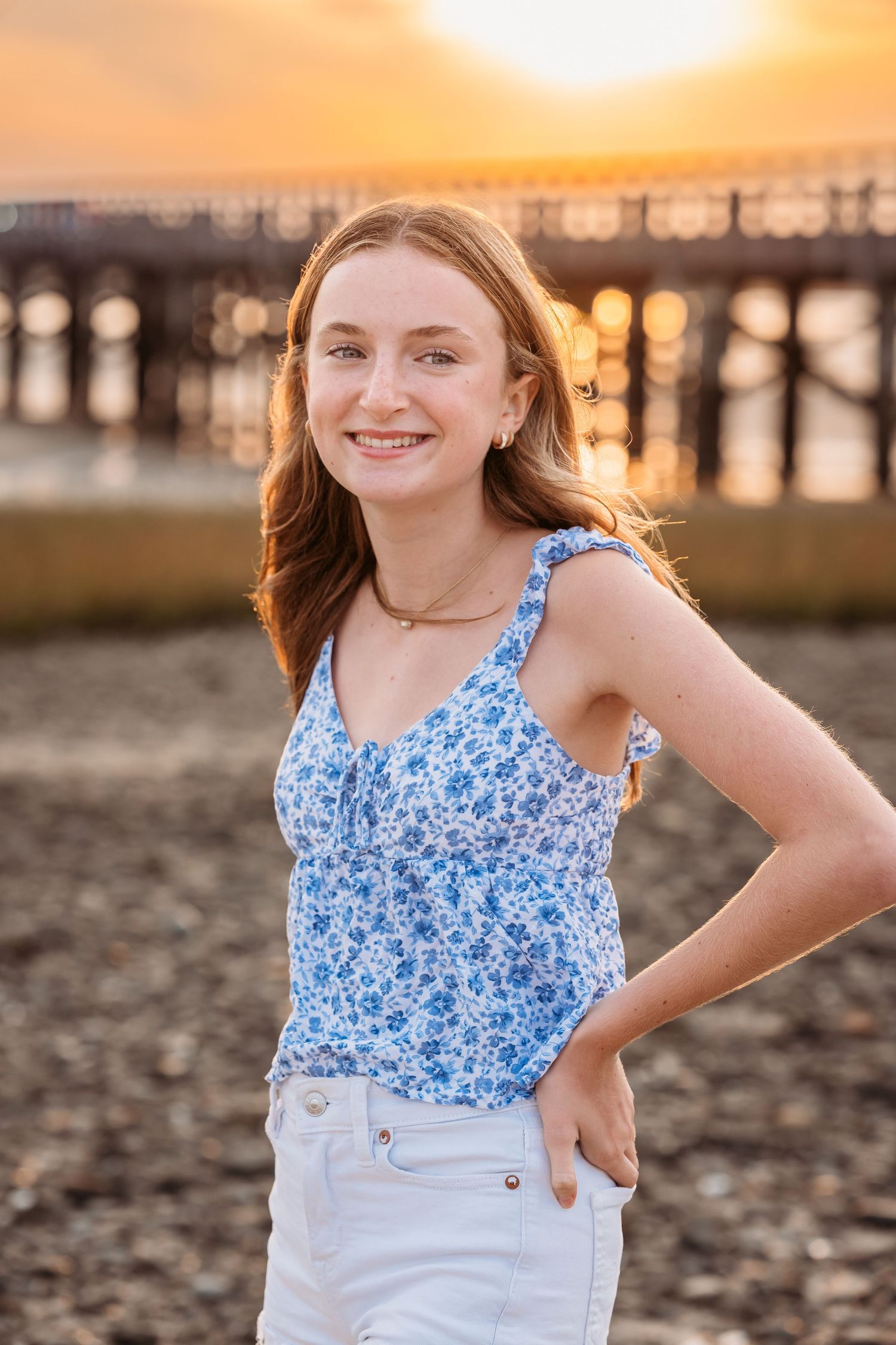 A person in a blue floral tank top and white shorts poses on the beach at sunset with a pier in the background.