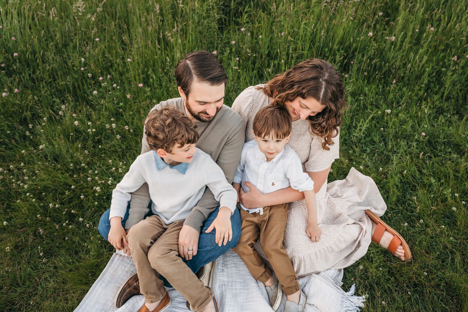 Family sits together on white blanket in tall grass during outdoor photo session.