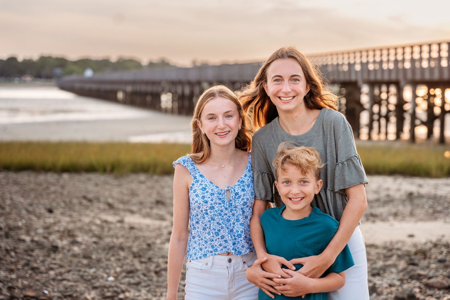 Family smiling together on a beach at sunset with wooden pier and marsh grass in background.