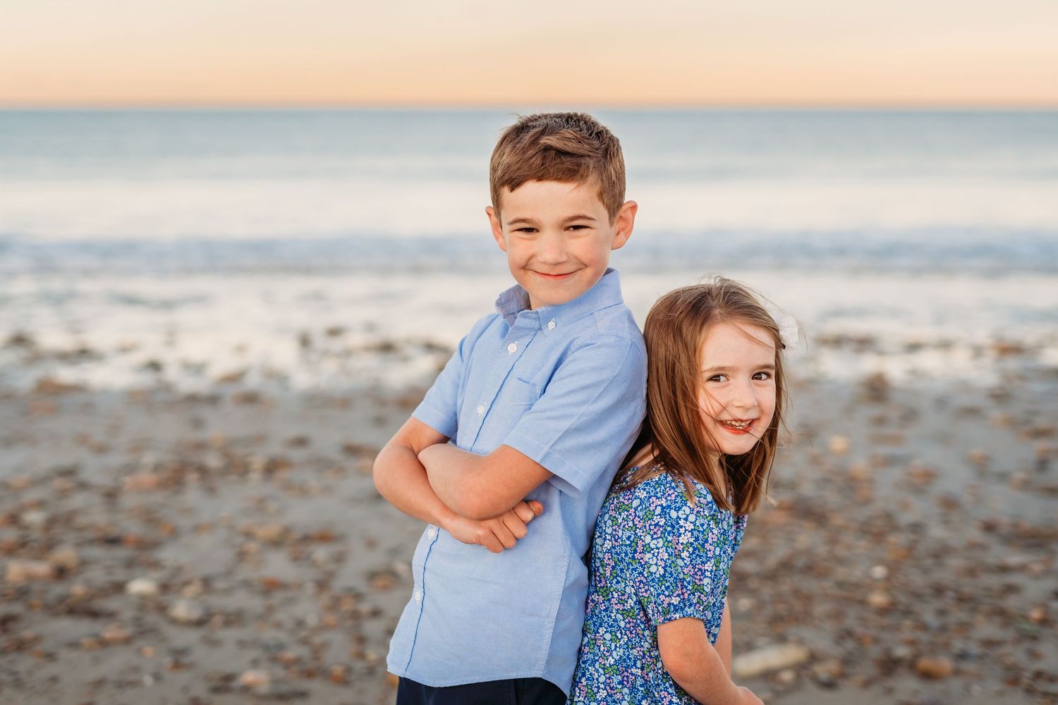 Two children stand back to back on a beach at sunset, smiling and posing for the camera.