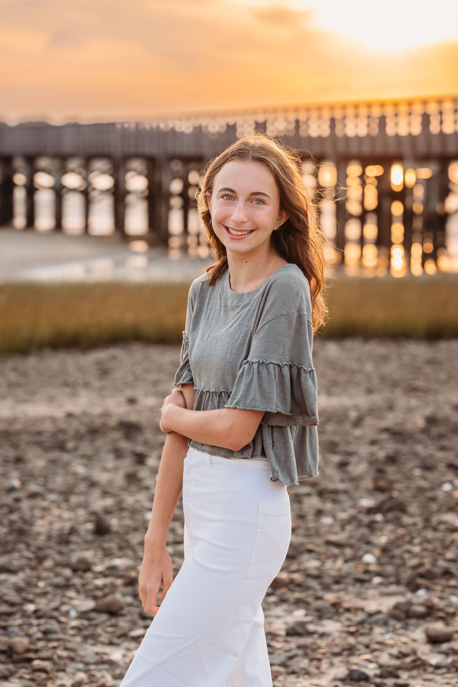 Person in gray ruffled top and white skirt poses on beach at sunset with wooden pier in background.