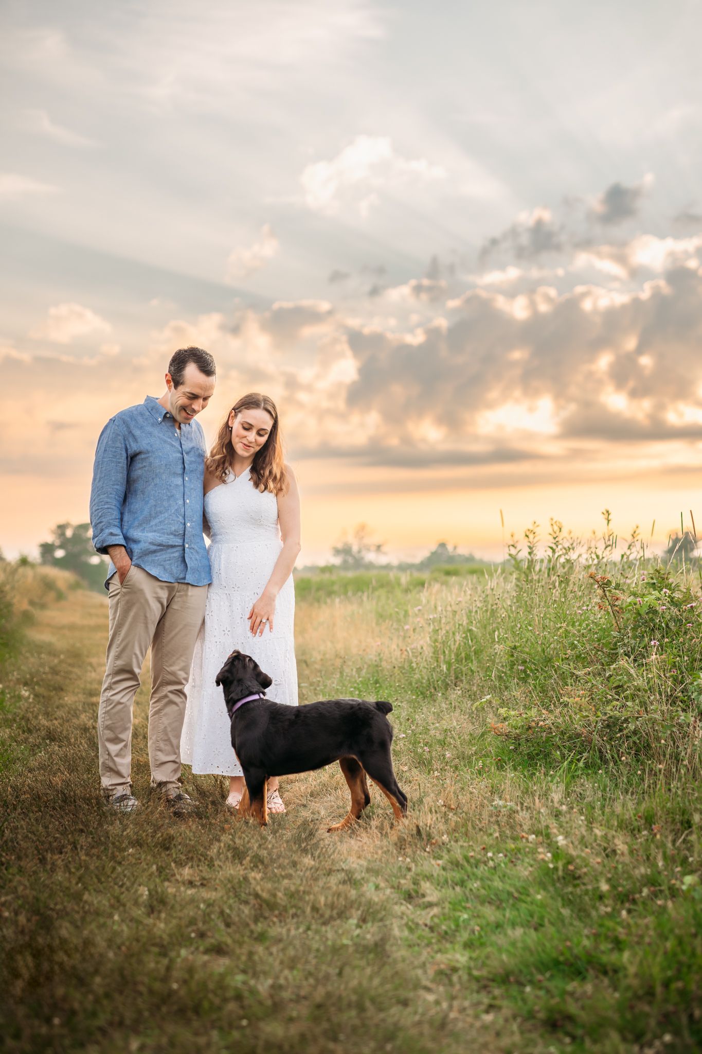 Couple stands with their black dog in a meadow during golden hour sunset.