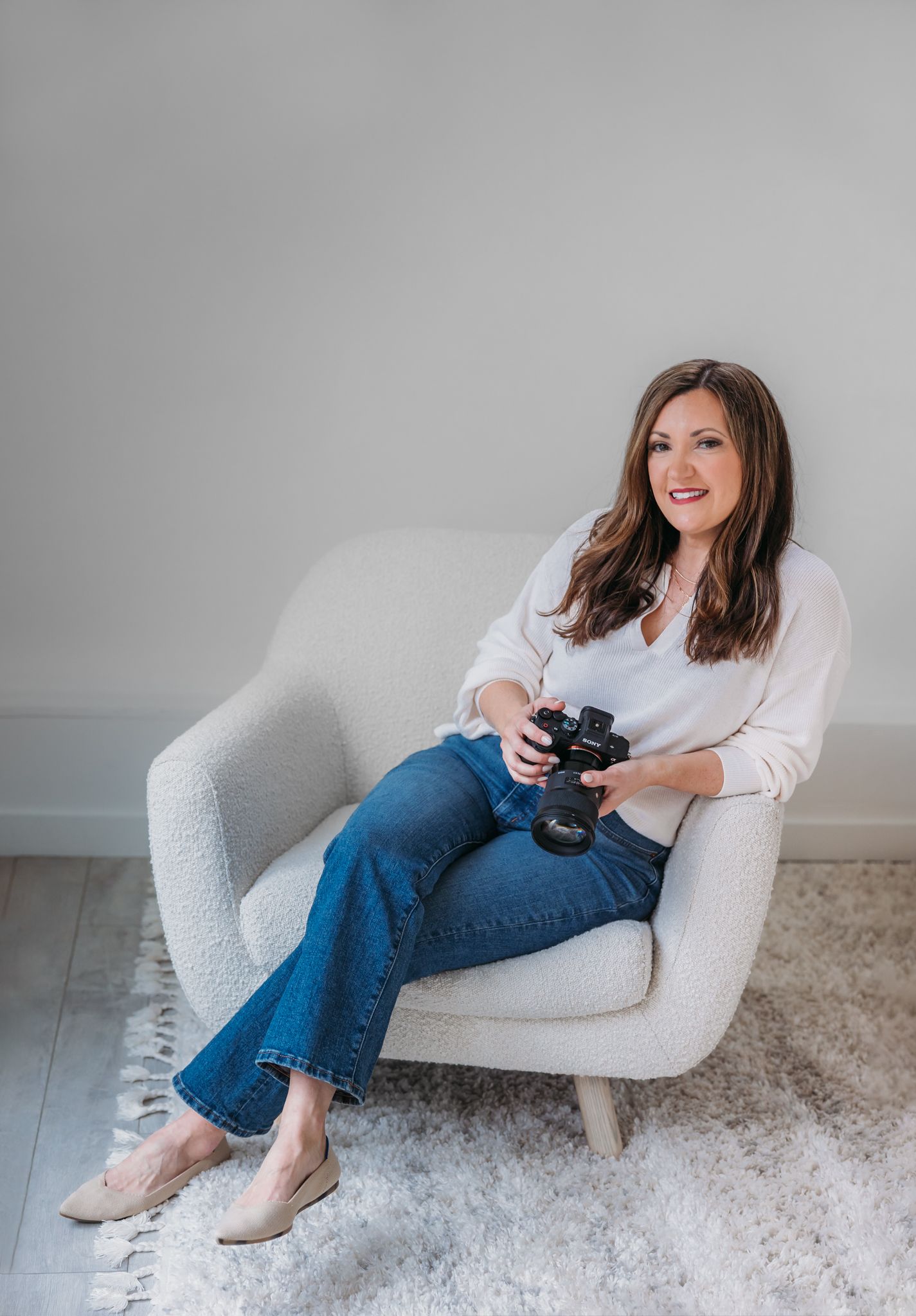 Professional photographer sits in a modern gray armchair holding a camera.