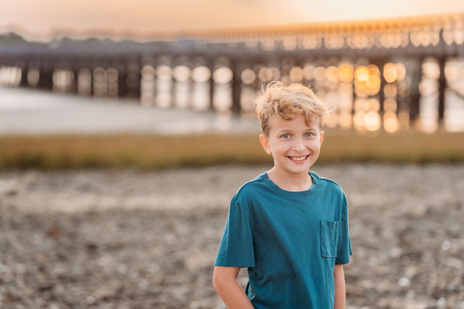 Young child in teal t-shirt smiling on beach with pier and sunset in background.