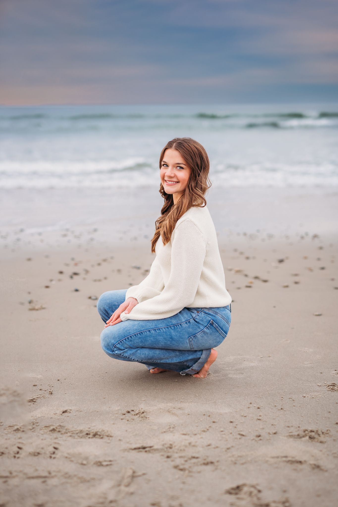 Candid lifestyle shots of someone in jeans and cream sweater sitting on sandy beach at sunset with ocean waves behind.