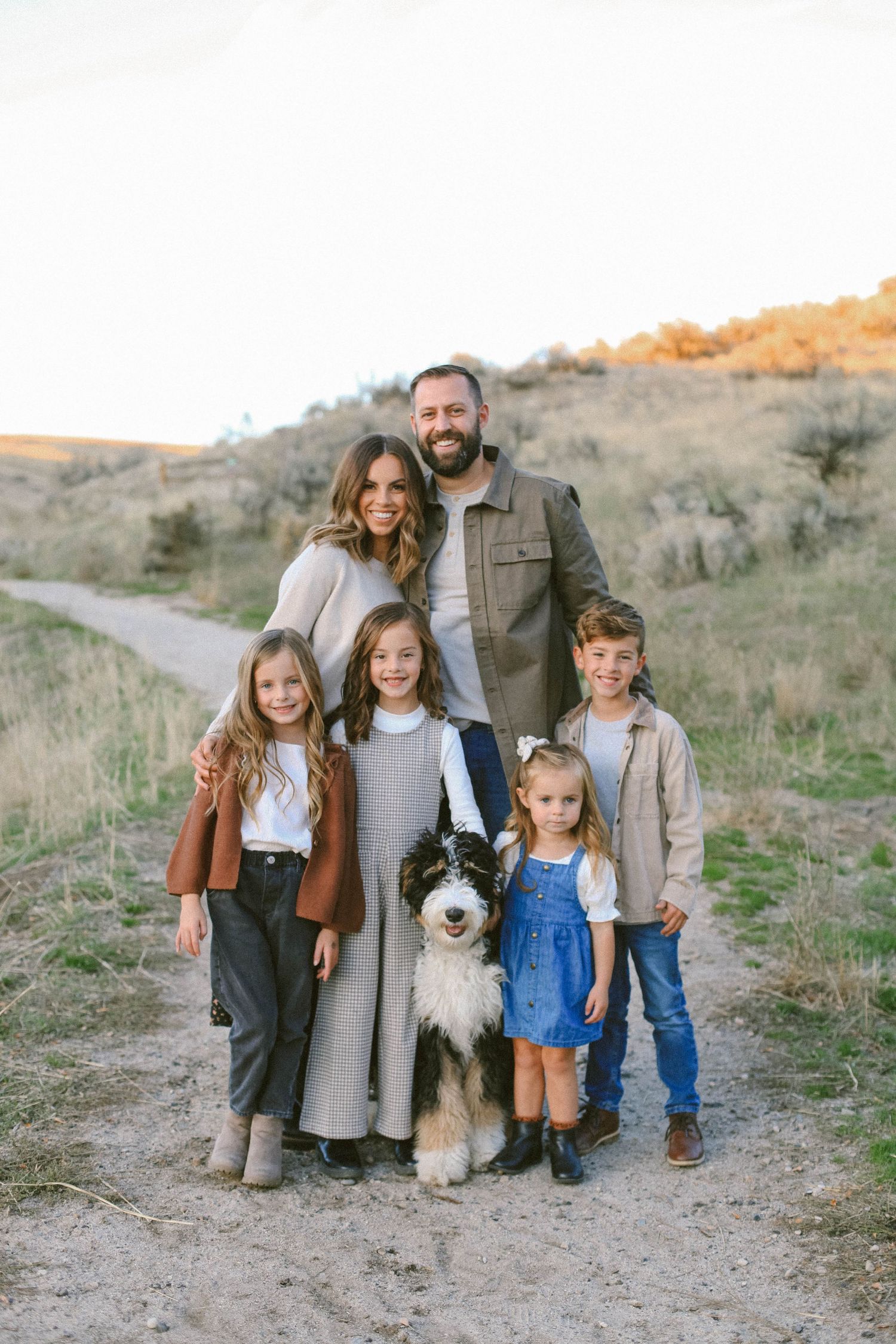 A family with children and their Australian Shepherd dog pose together on a dirt path in an autumn landscape.