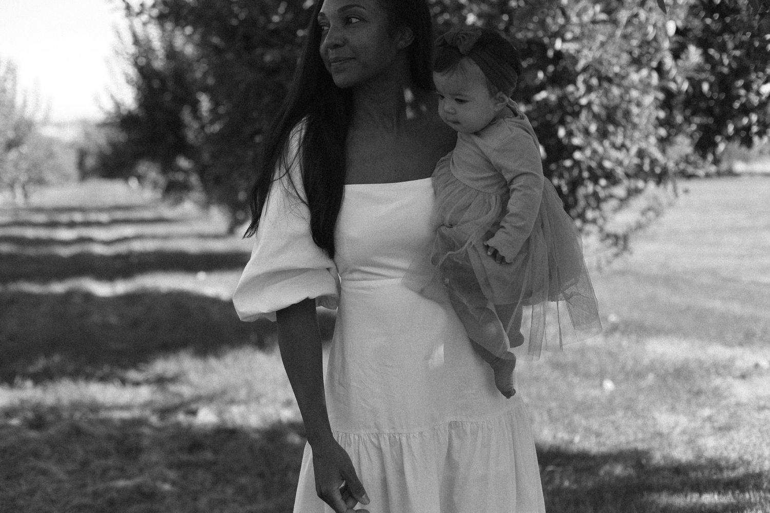 Woman in white dress holds a toddler while standing in a sunlit garden path.