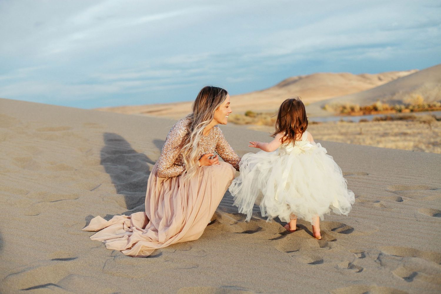 Mother and daughter in flowing dresses sitting together on sand dunes at sunset.