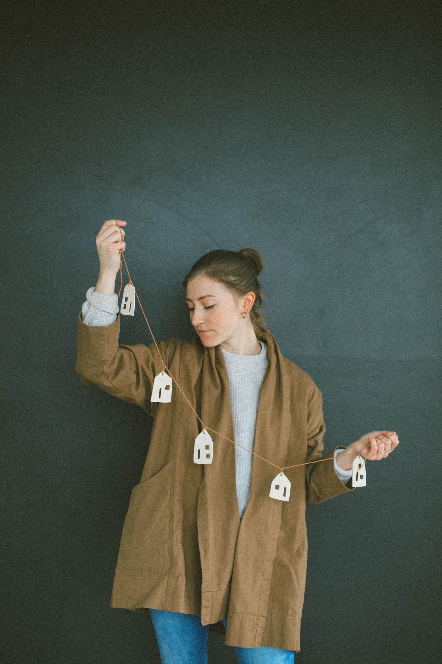 Women in brown jacket holding paper house cutouts against dark background in artistic pose.