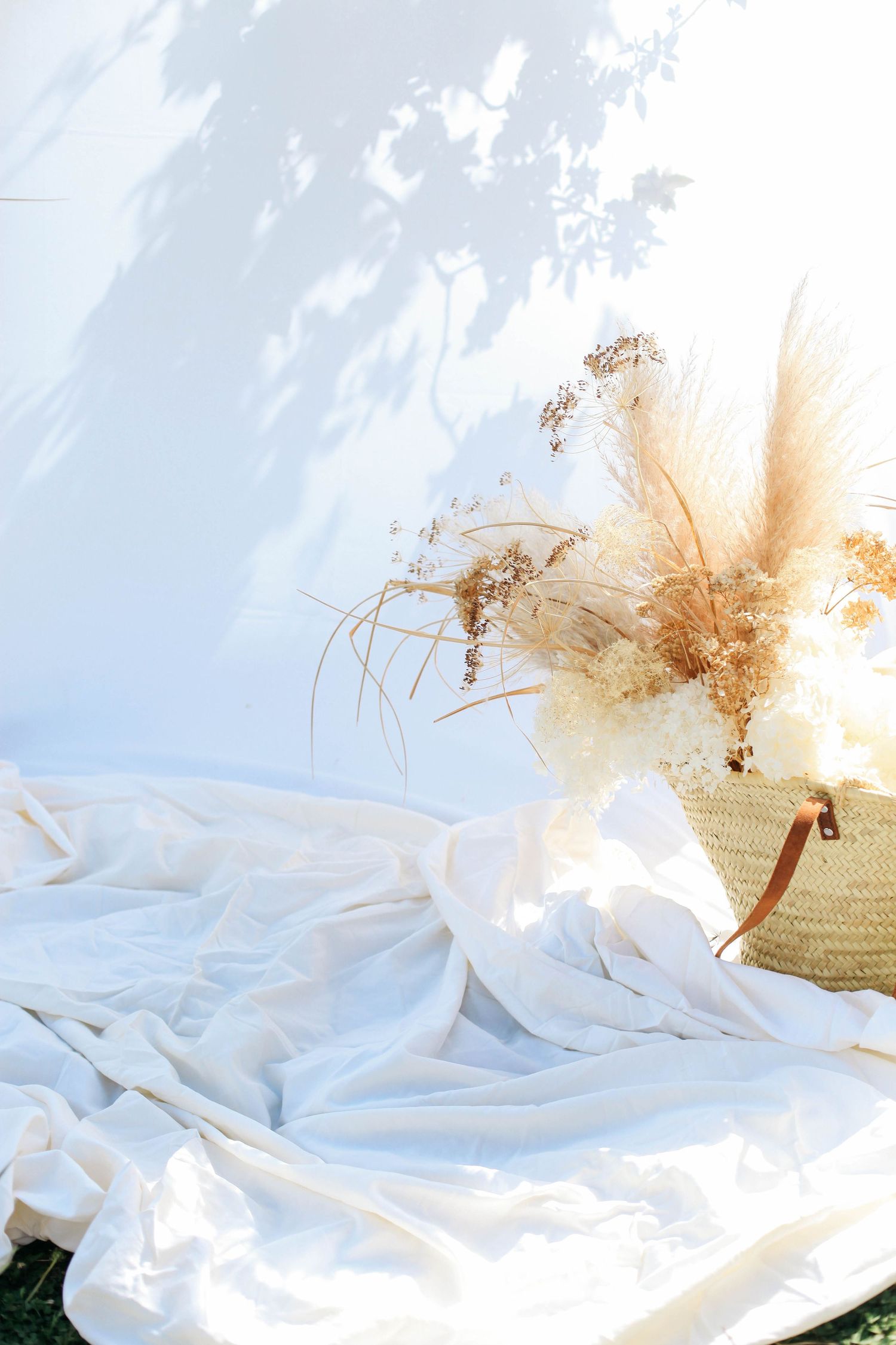 Ethereal still life with white fabric and dried flowers in straw basket against sky background.