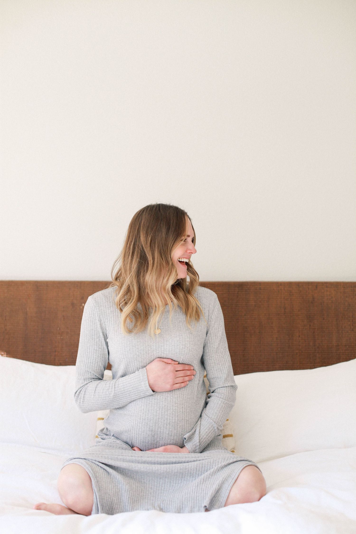 A pregnant woman in a gray sweater dress sits on a white bed looking to the side.