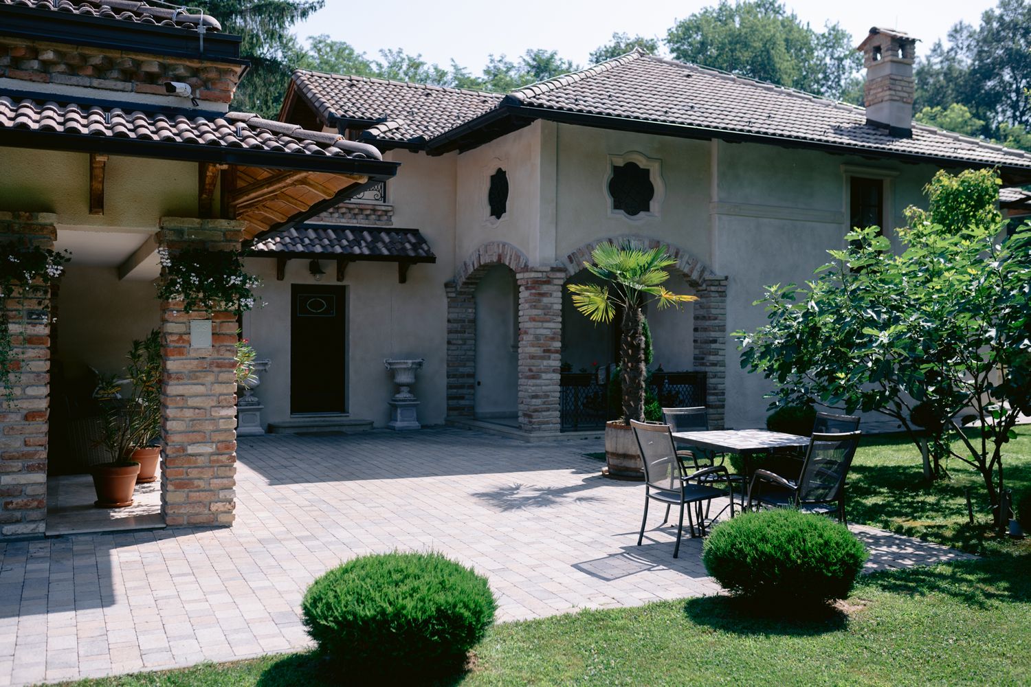 Mediterranean style patio with stone columns, tiled roof, and manicured garden featuring outdoor furniture and topiary.