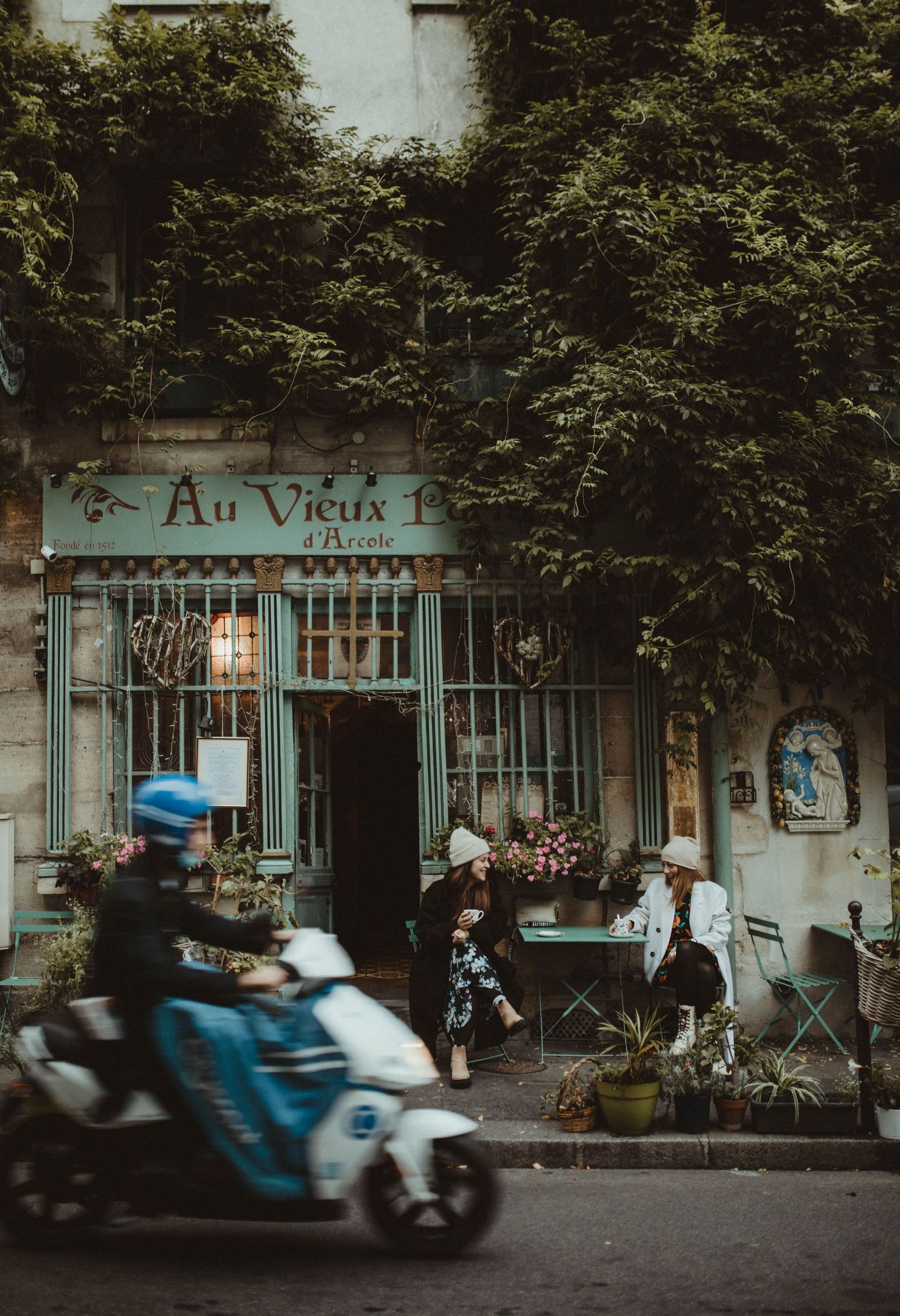 Quaint French cafe 'Au Vieux' with lush greenery and a passing blue scooter on a charming Parisian street.