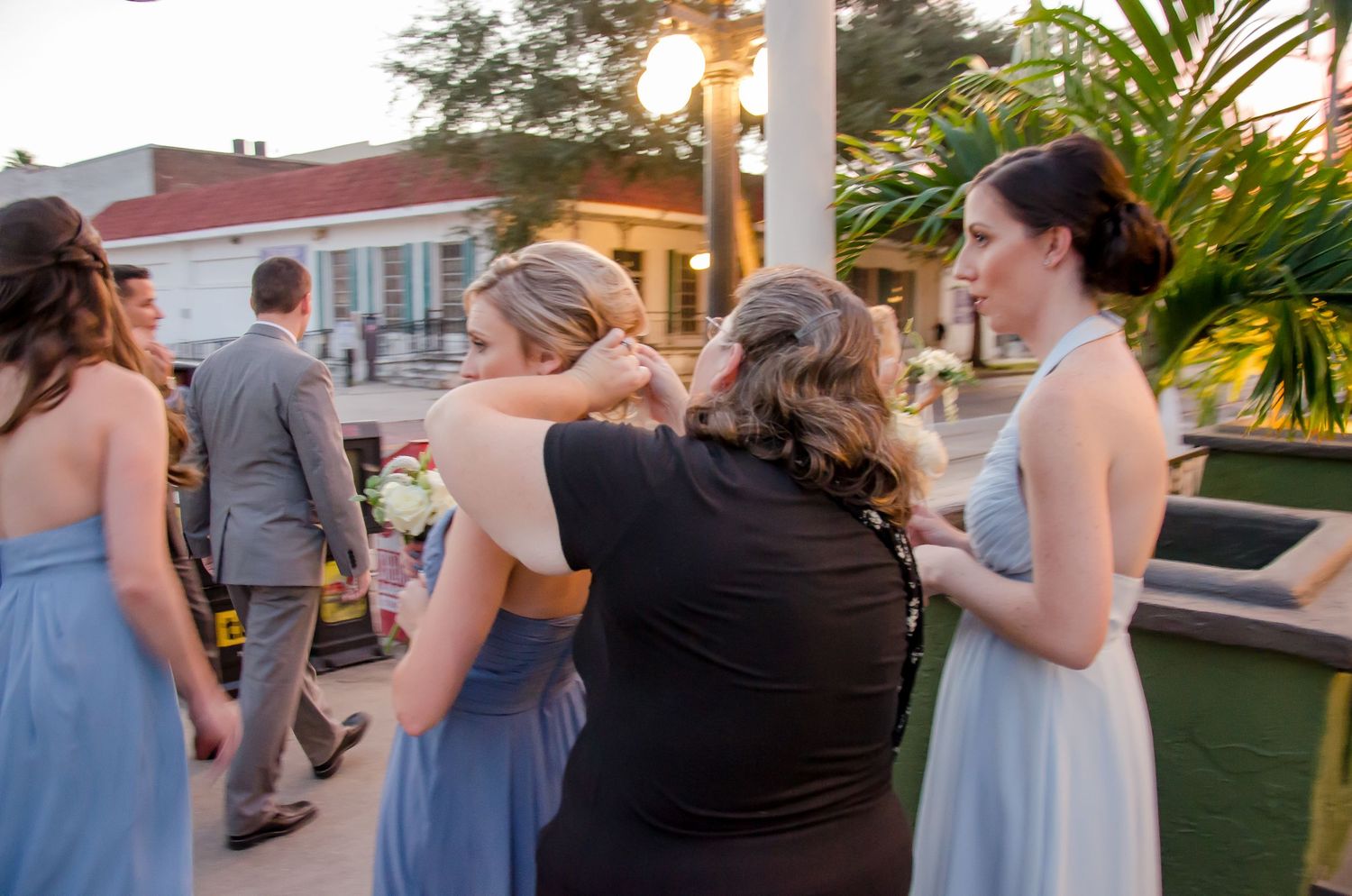 Wedding party guests in light blue dresses gather on an outdoor patio at sunset near tropical palm trees and lights.
