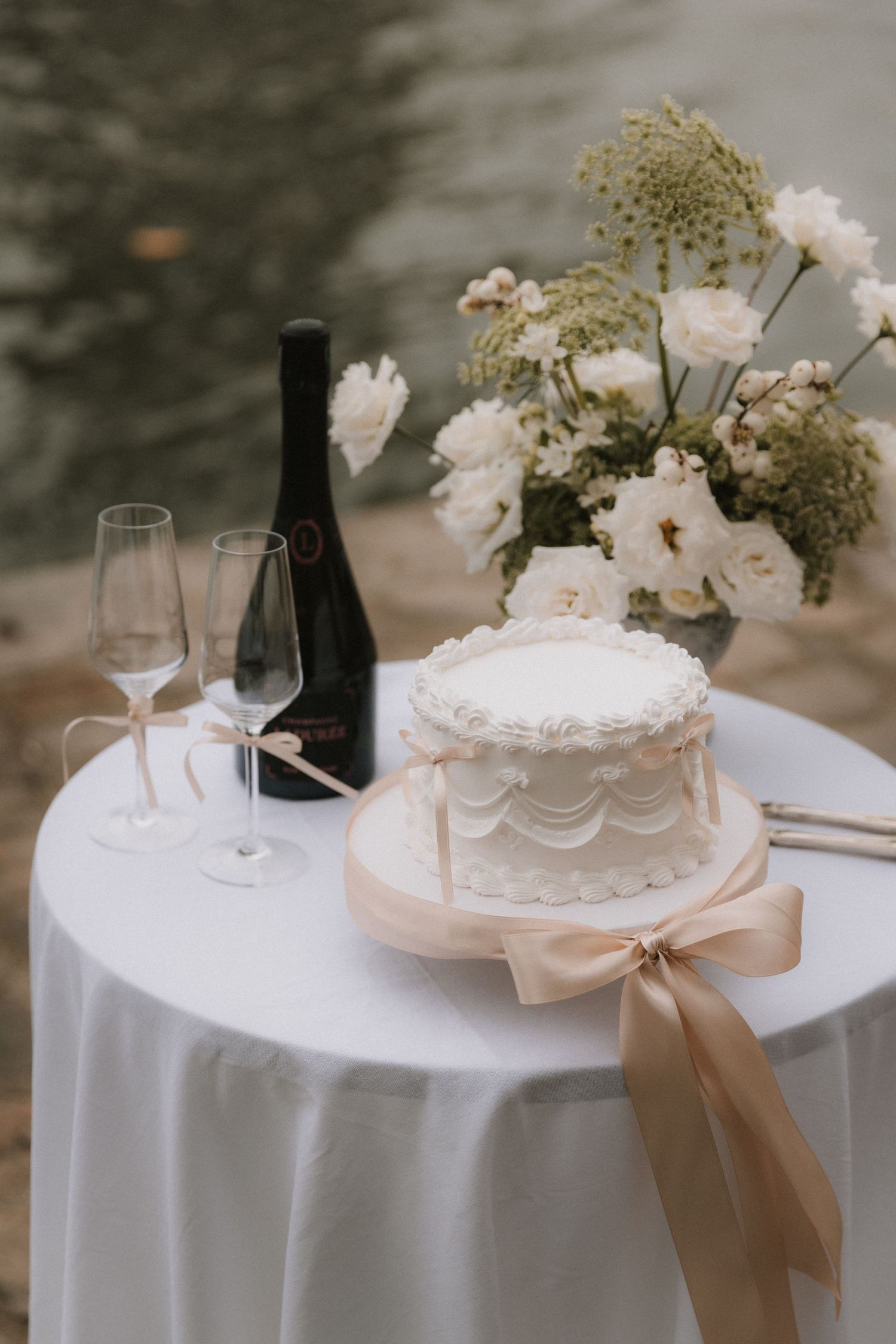 Elegant white wedding cake with champagne and floral arrangement on round table draped with white linen.