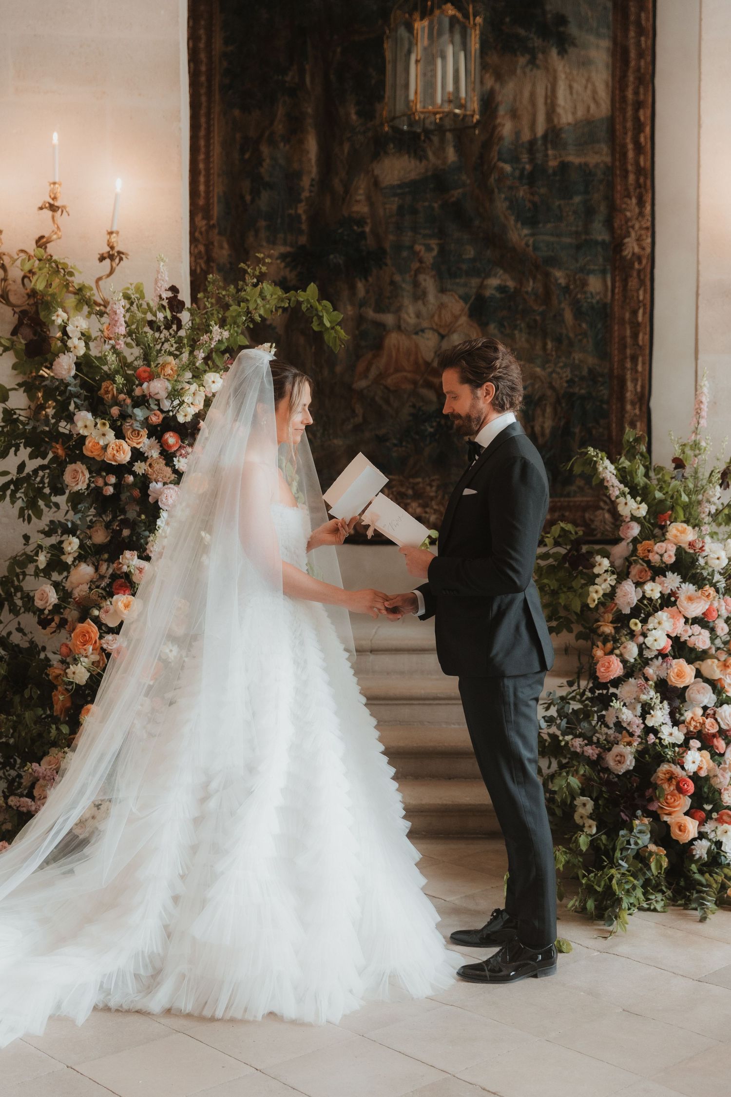 Couple exchanging vows in elegant room with stunning floral arrangements and tapestry backdrop during wedding ceremony.